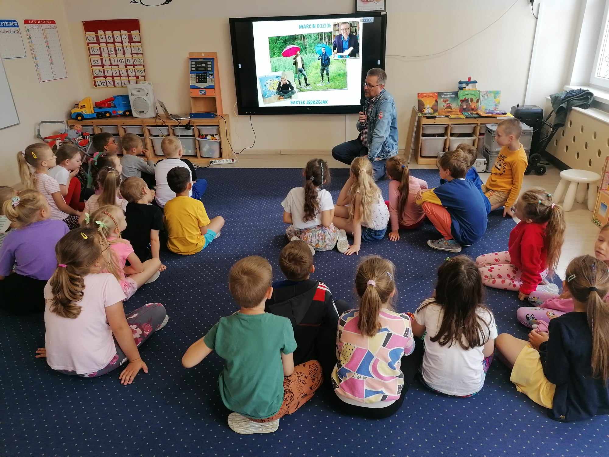 Autor książek z grupą dzieci przed tablicą multimedialną.